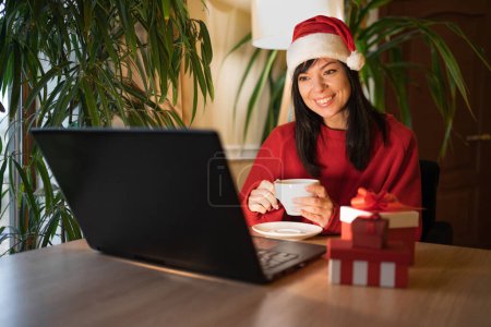 Photo for Young smiling woman watching movie on laptop smiling drinking tea in the evening on christmas eve. Copy space - Royalty Free Image