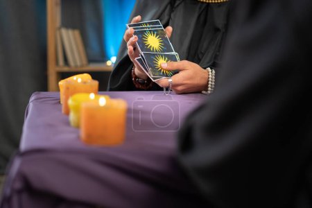 Photo for Close-up of a fortune tellers hand holding tarot cards over a table with candles, a future prediction session for a client. Copy space - Royalty Free Image