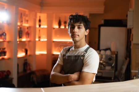 Photo for Handsome young bartender in apron looking at camera while standing at the bar counter with arms crossed. Copy space - Royalty Free Image