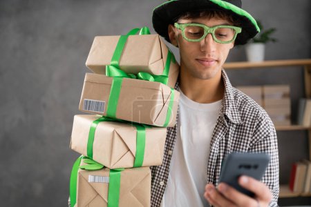 St. patrick's day sale, online shopping man in green leprechaun hat with stack of gift boxes using smartphone. Portrait