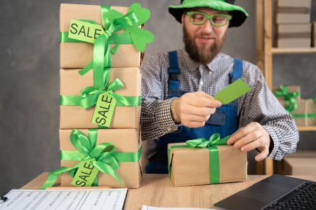 Téléchargez les photos : Small business owner with laptop and gift boxes with sale tags on table in home warehouse. Online ecommerce retail business St.Patricks Day discounts deals, free shipping. Best buy holiday offers - en image libre de droit