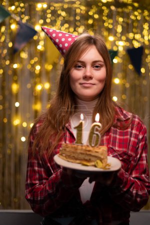 Photo for Birthday celebration. Portrait of cheerful young woman blow up candles 16 on birthday cake wears cone party hat makes wish have happy festive mood celebrated birthday at home. Festivity concept - Royalty Free Image