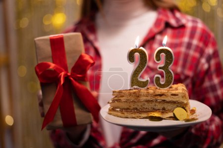 Téléchargez les photos : Young woman holding plate with tasty birthday cake and gift against defocused lights. 23th birthday. Copy space - en image libre de droit