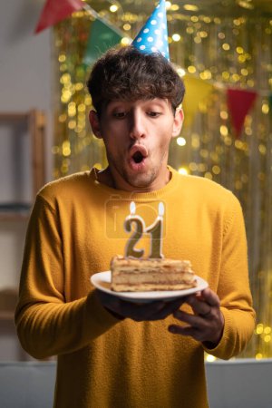 Photo for Holidays and celebration. Excited young man celebrating 21th birthday, blowing candles on cake, wearing party hat and having fun, standing over decorated home background. Copy space - Royalty Free Image