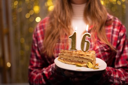 Young woman holding plate with tasty birthday cake with 16 number candle against defocused lights. Copy space
