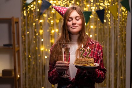 Birthday celebration. Caucasian girl makes a wish closing her eyes holding a birthday cake with candles number 22 in her hands. Copy space