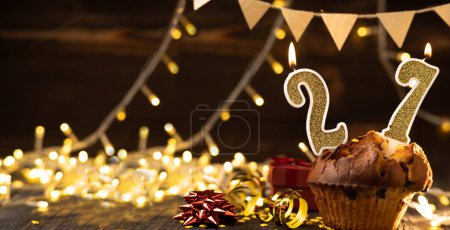 Foto de Number 27 gold burning candle in a cupcake against celebration wooden background with lights. Birthday cupcake. Copy space. Banner - Imagen libre de derechos