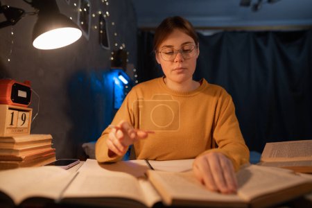 Focused student surrounded by books studying in dormitory at night. Copy space