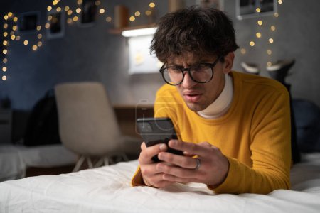 Photo for Upset confused young man holding cellphone having problem with mobile phone or missed call, frustrated angry guy student reading bad news in message looking at smartphone annoyed by spam. Copy space - Royalty Free Image