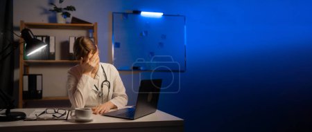 Photo for Tired woman doctor sitting at table and holding head in clinic during night shift. Banner. Copy space - Royalty Free Image