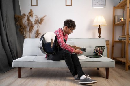Photo for Young man on sofa playing the guitar using laptop for online lesson at home. Copy space - Royalty Free Image