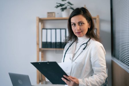 Photo for Portrait of smiling female doctor with lab coat in her office holding a clipboard with medical records looking at camera. Copy space - Royalty Free Image