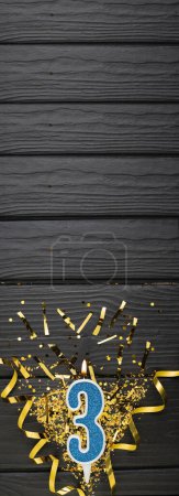 Téléchargez les photos : Number 3 blue celebration candle and gold confetti on dark wooden background. 3rd birthday card. Anniversary and birthday concept. Vertical banner. Copy space - en image libre de droit