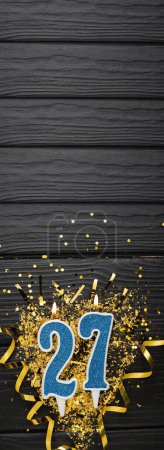 Téléchargez les photos : Number 27 blue celebration candle and gold confetti on dark wooden background. 27th birthday card. Anniversary and birthday concept. Vertical banner. Copy space - en image libre de droit