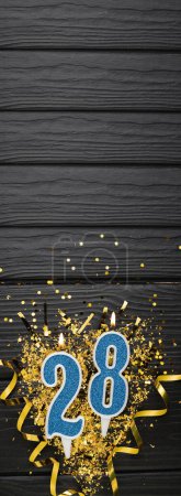 Téléchargez les photos : Number 28 blue celebration candle and gold confetti on dark wooden background. 28th birthday card. Anniversary and birthday concept. Vertical banner. Copy space - en image libre de droit