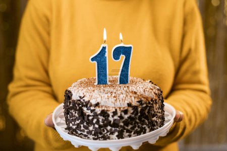 Photo for Woman holding birthday cake with burning candles number 17 against decorated background, close up. Celebrates birthday holiday at home. Copy space - Royalty Free Image