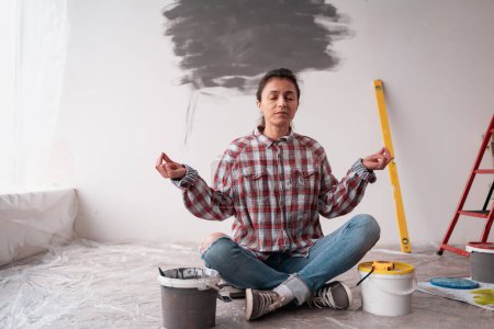 Téléchargez les photos : Femme heureuse assise sur le sol méditant, faisant du yoga et relaxant, salle d'appartement de rénovation. Outils de peinture murale. Réparation concept de maison - en image libre de droit