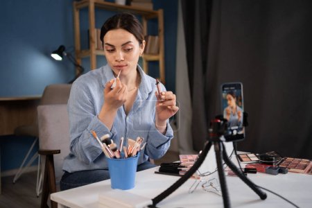 Photo for Beauty blogger caucasian young woman is broadcasting and recording the broadcast on a smartphone. applying lipstick on her lips and browsing beauty product in makeup vlogger. - Royalty Free Image