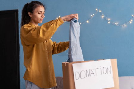 Photo for Concept of volunteering work and donation. Young woman helping poor people, packing cardboard donation box with clothes. Copy space - Royalty Free Image