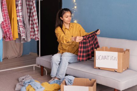 Photo for Woman sorting clothes sitting on sofa at home, packing cardboard donation box. Helping poor people. Concept of volunteering work, donation and clothes recycling. - Royalty Free Image
