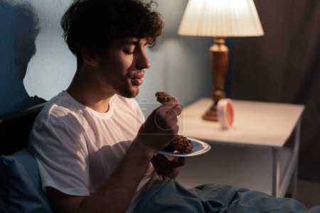 Foto de Joven estresado comiendo galletas por la noche. Persona deprimida cansada e infeliz comiendo durante la noche. Concepto de insomnio - Imagen libre de derechos