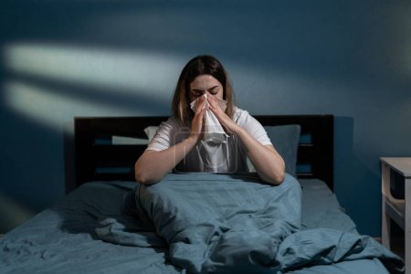 Photo for Sick young woman in bed blowing nose in napkin at night. Lady suffering cold and flu having temperature sitting on bed. Copy space - Royalty Free Image