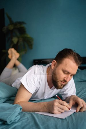 Photo for Handsome man laying on bed and writing a diary using notebook and pen. Copy space - Royalty Free Image