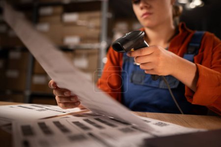 Téléchargez les photos : Gros plan de l'employé de l'entrepôt scannant les codes à barres sur du papier travaillant dans un grand entrepôt, assis à table. Espace de copie - en image libre de droit