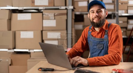Téléchargez les photos : Portrait d'un jeune travailleur masculin heureux travaillant sur un ordinateur portable dans un entrepôt, Préparation de colis pour la livraison. Espace de copie - en image libre de droit