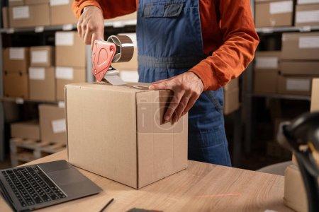 Worker man In Retail Warehouse working packing parcel box with tape dispenser for shipment. Delivery and distribution center full of shelves with goods. Close-up. Copy space