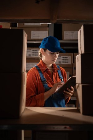 Téléchargez les photos : Gestionnaire vérification des stocks utilise tablette numérique. Jeune femme travaillant dans un entrepôt avec des rangées d'étagères pleines de colis. Espace de copie - en image libre de droit