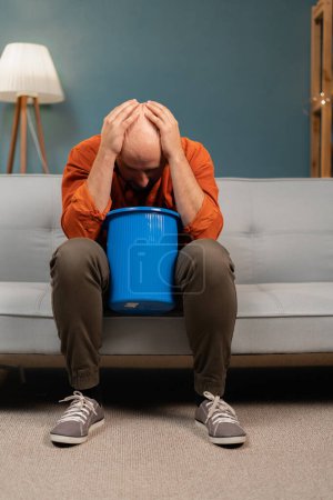 Photo for Bearded man vomit into bucket sitting on couch at home. Intoxication, poisoning and nausea concept - Royalty Free Image