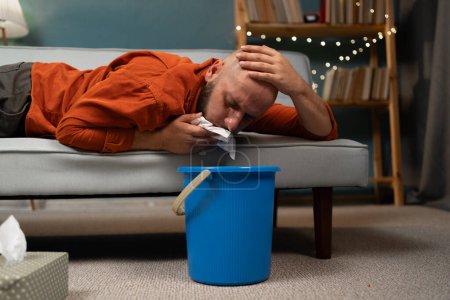 Photo for Man vomiting into a bucket on couch at home. Intoxication, poisoning and nausea concept. Close-up - Royalty Free Image