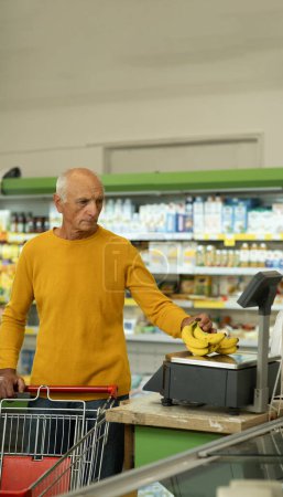 Photo for Elderly man weighing bananas on scales at supermarket. Senor shopping for food, buying fruits at grocery store. Copy space - Royalty Free Image