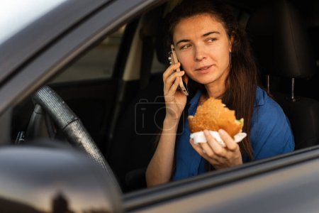 Téléchargez les photos : Femme mangeant un hamburger et parlant au téléphone alors qu'elle était assise dans une voiture. Restauration rapide et à emporter sur la route. Espace de copie - en image libre de droit