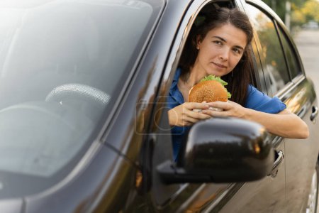 Téléchargez les photos : Femme conducteur mangeant un délicieux hamburger gras dans sa voiture tout en profitant du déjeuner tout en se tenant dans un embouteillage. Espace de copie - en image libre de droit