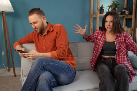 Téléchargez les photos : Agacé jeune femme en colère criant sur son homme qui utilise un smartphone et en ignorant sa femme confus. Conflit relationnel concept. Espace de copie - en image libre de droit