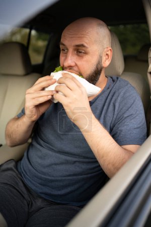 Téléchargez les photos : Homme mangeant hamburger en voiture tout en prenant la pause déjeuner. Espace de copie - en image libre de droit