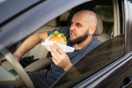 Téléchargez les photos : Homme fatigué conduisant une voiture tout en mangeant un hamburger. En colère d'attendre debout dans un embouteillage. Espace de copie - en image libre de droit