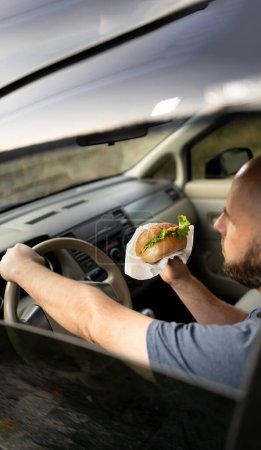 Téléchargez les photos : Homme conduisant une voiture en mangeant un hamburger debout dans un embouteillage le matin. Espace de copie - en image libre de droit