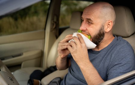 Téléchargez les photos : Homme heureux chauffeur manger un hamburger frais tout en conduisant une voiture. Le concept de commande de restauration rapide et de conduite dangereuse. Espace de copie - en image libre de droit