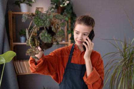 Photo for Smiling child girl gardener talking by mobile phone in workshop transplantation houseplants. Planting of home green plants in home garden, hobby. Copy space - Royalty Free Image