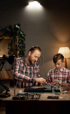 Téléchargez les photos : Père avec fils en utilisant du fer à souder pour réparer l'ordinateur portable à la maison. Concept d'entretien et de famille. Espace de copie - en image libre de droit