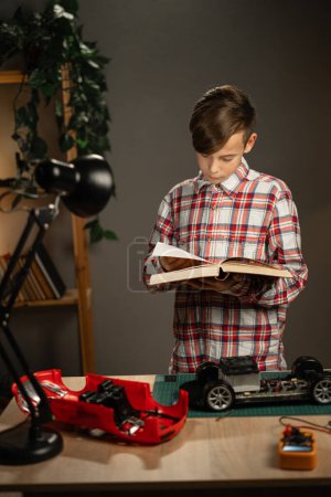 Téléchargez les photos : Garçon concentré lisant le livre de science et utilisant le fer à souder pour fixer la voiture jouet télécommandée à la maison. Espace de copie - en image libre de droit