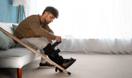 Hombre lesionado con bota de caminante u ortesis de tobillo sentado en el sofá en casa, espacio para copiar.