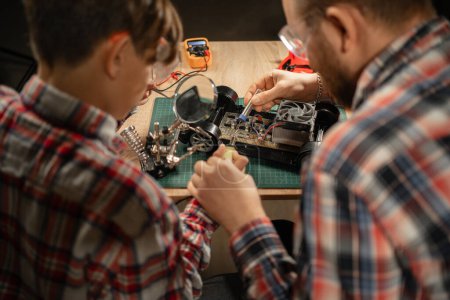 Photo for Father and son in safety glasses are soldering a broken machine on the control panel, view over the shoulder. Close-up - Royalty Free Image