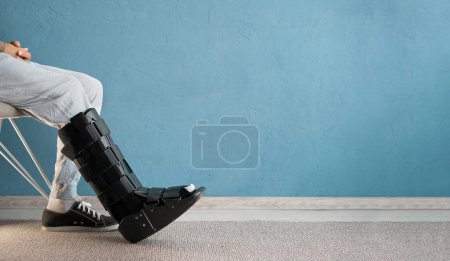 Photo for Injuried man with orthosis and sprained ankle sitting on the chair. Ankle orthosis concept. Copy space. Banner - Royalty Free Image