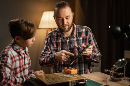 Photo for Caucasian father and his son using soldering iron for fixing motherboard from laptop. Family sitting together at table and repairing computer at home. Copy space - Royalty Free Image