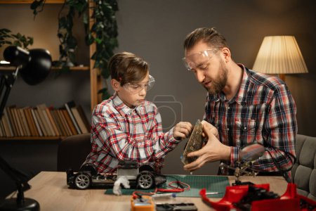 Photo for Father and son in protective glasses using soldering iron for repairing toy car at home in the evening. Concept of family and togetherness. Copy space - Royalty Free Image