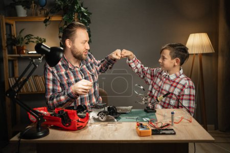 Téléchargez les photos : Carte de circuit imprimé à souder père et fils avec fer à souder à la maison se réjouissant en cognant leurs poings comme un signe d'amitié. Espace de copie - en image libre de droit